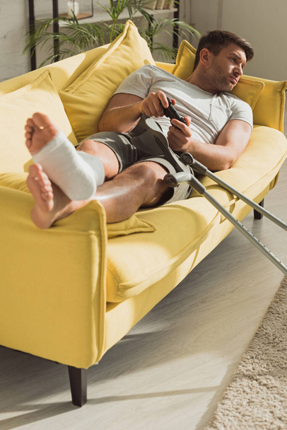 KYIV, UKRAINE - JANUARY 21, 2020: Selective focus of man with broken leg playing video game on sofa at home - Fotoğraf, Görsel