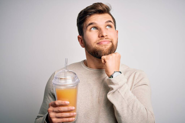 Young blond man with beard and blue eyes drinking healthy orange smoothie serious face thinking about question, very confused idea - Photo, image
