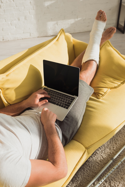 Cropped view of man with broken leg typing on laptop on sofa - 写真・画像