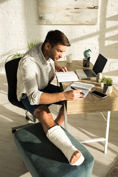 Side view of handsome freelancer with broken leg working with notebooks and laptop at table - Photo, Image