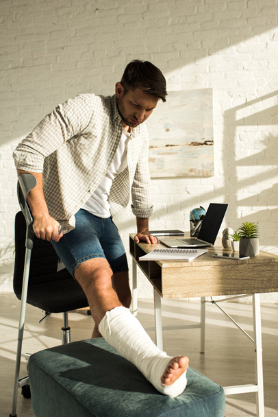 Man with broken leg standing near table with laptop and notebooks - 写真・画像