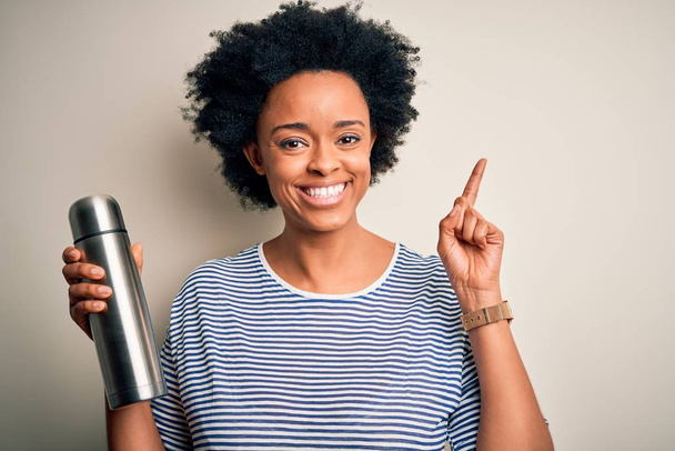 Jonge Afro-Amerikaanse afro toeristische vrouw met krullend haar houden thermo met water verrast met een idee of vraag wijzend vinger met gelukkig gezicht, nummer een - Foto, afbeelding