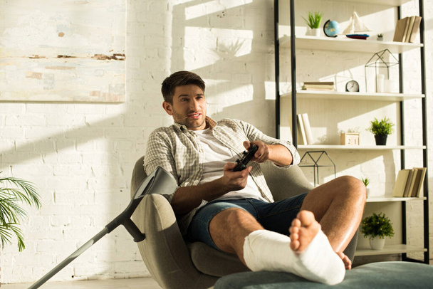 KYIV, UKRAINE - JANUARY 21, 2020: Handsome man with broken leg on ottoman playing video game at home - Photo, Image