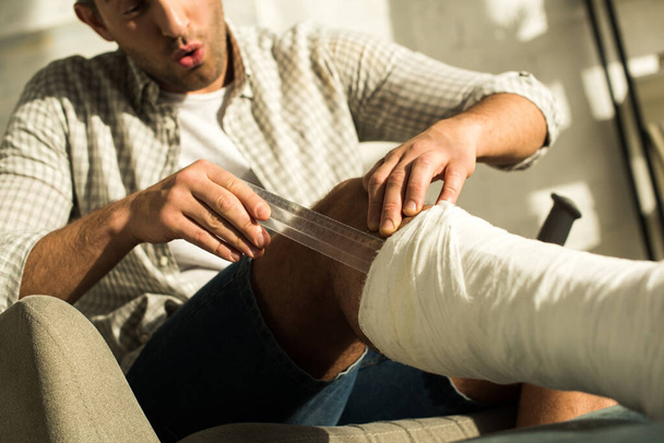Foco seletivo do homem saído coçando a perna em bandagem de gesso com régua na poltrona
 - Foto, Imagem