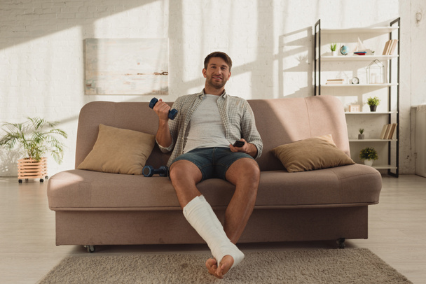 Mosolygós férfi törött lábbal néz TV-t és tartja súlyzó kanapén otthon - Fotó, kép