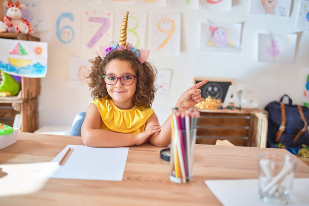 Beautiful toddler wearing glasses and unicorn diadem sitting on desk at kindergarten with a big smile on face, pointing with hand and finger to the side looking at the camera. - 写真・画像
