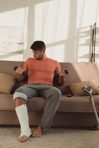 Hombre guapo con pierna rota entrenando con mancuernas en el sofá en casa
 - Foto, imagen