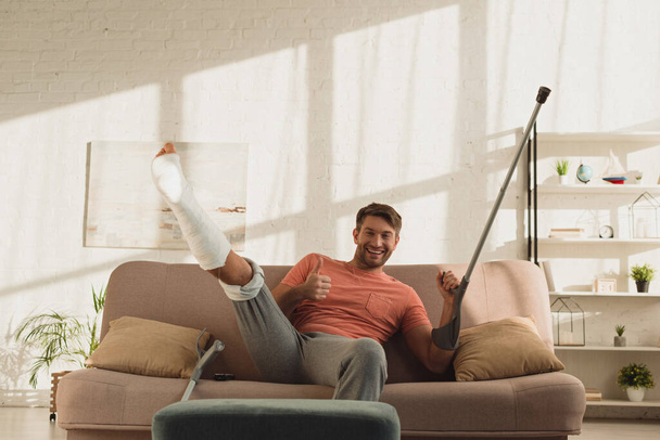 Hombre guapo con la pierna rota levantada y la muleta mostrando el cartel de aprobación en la cámara en el sofá
 - Foto, imagen