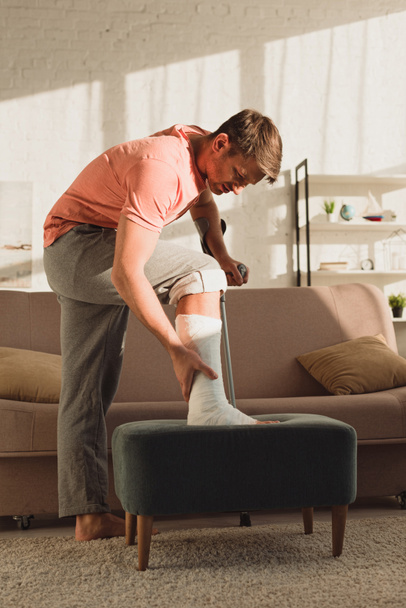 Вид збоку чоловіка торкається зламаної ноги на пуфеті вдома
 - Фото, зображення