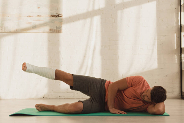 Man with leg in plaster bandage training on fitness mat on floor at home - Fotografie, Obrázek