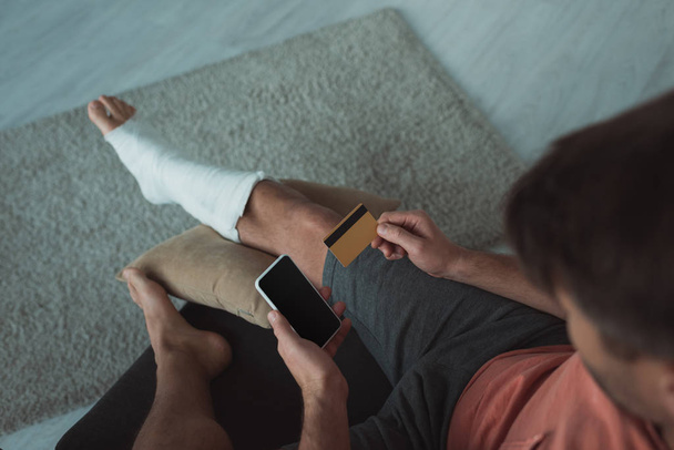 Widok na człowieka ze złamaną nogą przy użyciu smartfona i karty kredytowej na kanapie - Zdjęcie, obraz