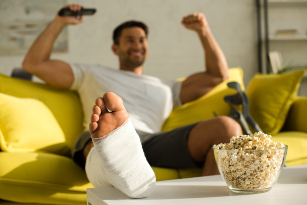 Selective focus of popcorn and broken leg of cheerful man with remote controller on couch - Photo, Image