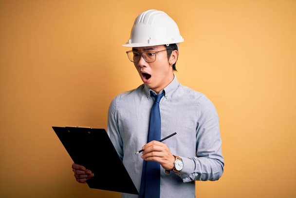 Young handsome chinese engineer man wearing safety helmet holding clipboard scared in shock with a surprise face, afraid and excited with fear expression - 写真・画像