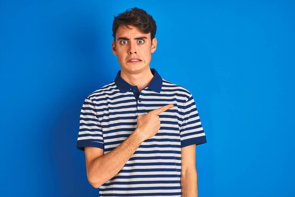 Teenager boy wearing casual t-shirt standing over blue isolated background Pointing aside worried and nervous with forefinger, concerned and surprised expression - Photo, Image