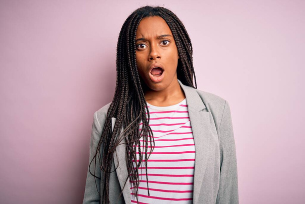 Jovem afro-americana empresária de pé sobre rosa isolado fundo em face de choque, olhando cético e sarcástico, surpreso com a boca aberta
 - Foto, Imagem