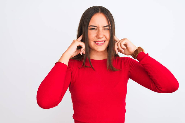 Jong mooi meisje draagt rode casual t-shirt staan over geïsoleerde witte achtergrond bedekt oren met vingers met geïrriteerde uitdrukking voor het lawaai van luide muziek. Dovemansconcept. - Foto, afbeelding