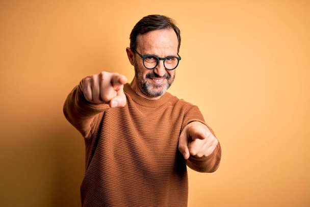 Middle age hoary man wearing brown sweater and glasses over isolated yellow background pointing to you and the camera with fingers, smiling positive and cheerful - Photo, Image
