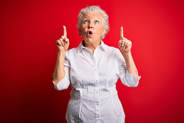 Старшая красивая женщина в элегантной рубашке стоя на изолированном красном фоне поражен и удивлен глядя вверх и указывая пальцами и поднятыми руками
. - Фото, изображение