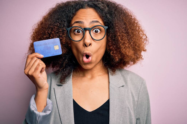 Jeune femme d'affaires afro-américaine avec des cheveux afro tenant carte de crédit sur fond rose effrayée en état de choc avec un visage surprise, effrayée et excitée par l'expression de la peur
 - Photo, image