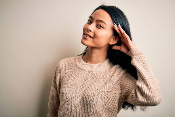 junge schöne Chinesin in lässigem Pullover vor isoliertem weißem Hintergrund, lächelnd mit der Hand über dem Ohr, um Gerüchten oder Klatsch zuzuhören. Konzept für Gehörlosigkeit. - Foto, Bild