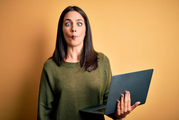 Fiatal barna nő kék szemekkel dolgozik számítógépes laptop sárga háttér, hogy hal arc ajkak, őrült és komikus gesztus. Vicces kifejezés.. - Fotó, kép