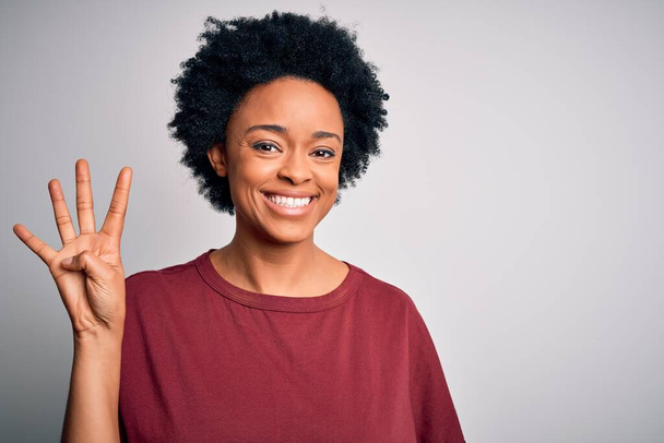 Młoda, piękna afro Afroamerykanka z kręconymi włosami, nosząca luźny t-shirt na stojąco, pokazująca i wskazująca palcami numer cztery, uśmiechnięta, pewna siebie i szczęśliwa.. - Zdjęcie, obraz