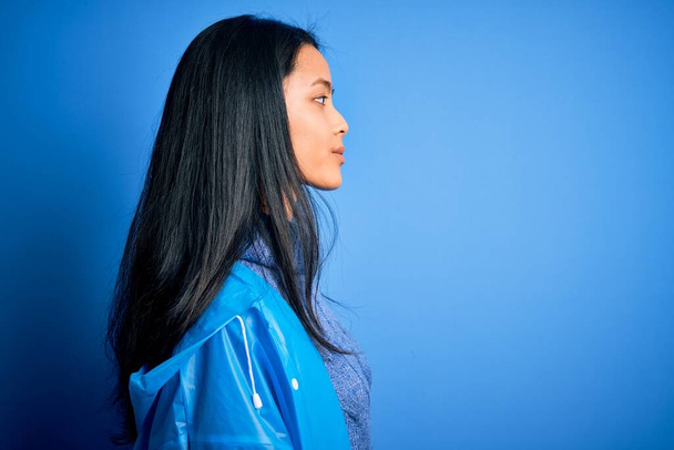 Młoda piękna Chinka nosi płaszcz przeciwdeszczowy stojąc na odizolowanym niebieskim tle patrząc na bok, relaks profil pozować z naturalną twarz z pewnym uśmiechem. - Zdjęcie, obraz
