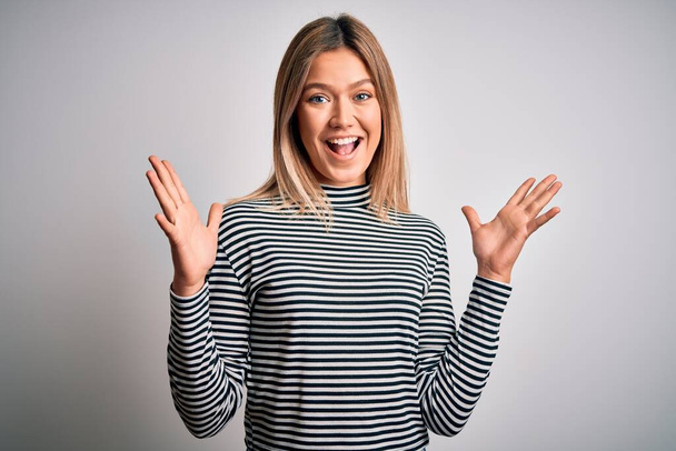 Young beautiful blonde woman wearing casual striped sweater over isolated background celebrating crazy and amazed for success with arms raised and open eyes screaming excited. Winner concept - Fotografie, Obrázek