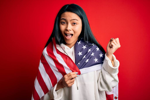 Jonge mooie Chinese vrouw dragen van de Verenigde Staten vlag over geïsoleerde rode achtergrond schreeuwen trots en vieren overwinning en succes zeer opgewonden, juichen emotie - Foto, afbeelding
