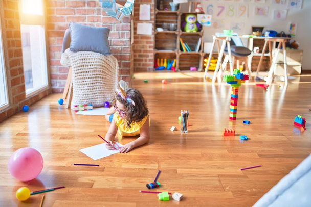 Hermoso niño pequeño con gafas y diadema de unicornio acostado en el dibujo del suelo con papel y lápiz en el jardín de infantes
 - Foto, imagen