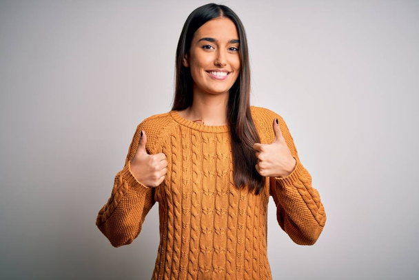 Young beautiful brunette woman wearing casual sweater over isolated white background success sign doing positive gesture with hand, thumbs up smiling and happy. Cheerful expression and winner gesture. - Foto, Imagem