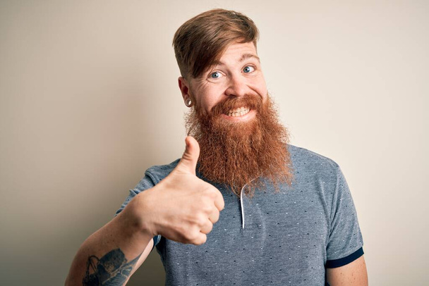Homme rousse irlandais beau avec barbe et tatouage de bras debout sur un fond isolé faisant geste heureux pouces vers le haut avec la main. Approuver l'expression en regardant la caméra montrant le succès
. - Photo, image