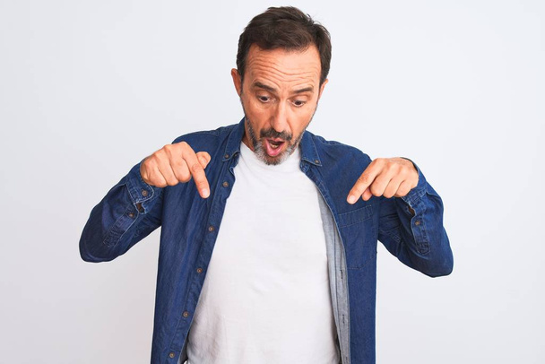 Мужчина средних лет в синей джинсовой рубашке стоит на изолированном белом фоне, указывая пальцами на рекламу, удивленное лицо и открытый рот.
 - Фото, изображение