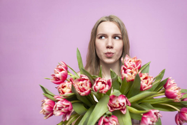 emotional face of a girl in flowers on a pink background, a surprised woman with a bouquet of tulips is smiling - Photo, Image