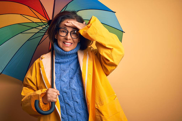 Kobieta w średnim wieku ubrana w żółty płaszcz przeciwdeszczowy pod kolorowym parasolem na odizolowanym tle, zestresowana dłonią na głowie, zszokowana wstydem i niespodziewaną twarzą, zła i sfrustrowana. Strach i zdenerwowanie przez pomyłkę. - Zdjęcie, obraz