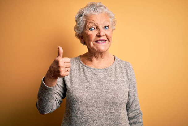 Senior belle femme portant t-shirt décontracté debout sur fond jaune isolé faire des pouces heureux geste avec la main. Approuver l'expression en regardant la caméra montrant le succès
. - Photo, image