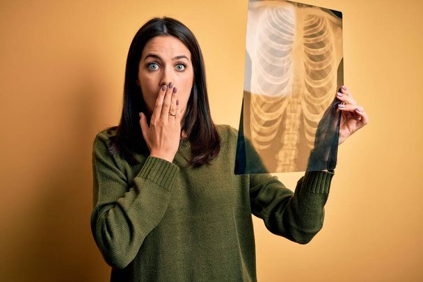 Young brunette woman with blue eyes holding xray of lungs over yellow background cover mouth with hand shocked with shame for mistake, expression of fear, scared in silence, secret concept - Fotoğraf, Görsel