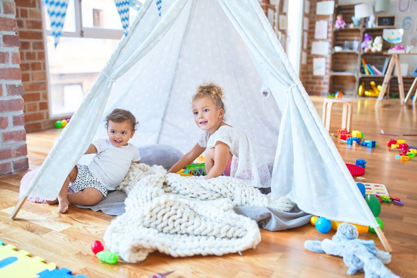 Hermosos niños jugando dentro de tipi sobre manta alrededor de un montón de juguetes en el jardín de infantes
 - Foto, Imagen