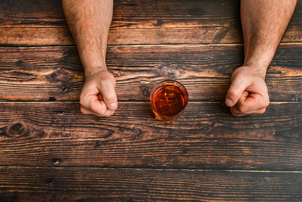 Ένας άντρας σφίγγει τις γροθιές του, συγκρατείται από το να πίνει αλκοόλ σε ένα ξύλινο τραπέζι. Η έννοια του αλκοολισμού και της εξάρτησης από το αλκοόλ. - Φωτογραφία, εικόνα