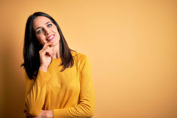 Giovane donna bruna con gli occhi azzurri che indossa maglione casual su sfondo giallo guardando fiducioso la fotocamera con il sorriso con le braccia incrociate e la mano sollevata sul mento. Pensare positivo
. - Foto, immagini