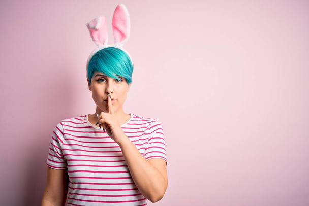 Молодая женщина с модными голубыми волосами, носящая пасхальные кроличьи уши на розовом фоне, просит тишины с пальцем на губах. Молчание и тайная концепция
. - Фото, изображение