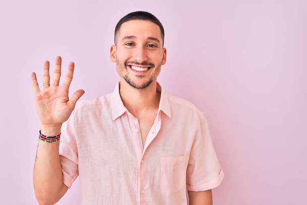 Fiatal jóképű férfi rózsaszín ingben áll elszigetelt háttér mutatja, és rámutatva ujjak száma 5 miközben mosolygós magabiztos és boldog. - Fotó, kép