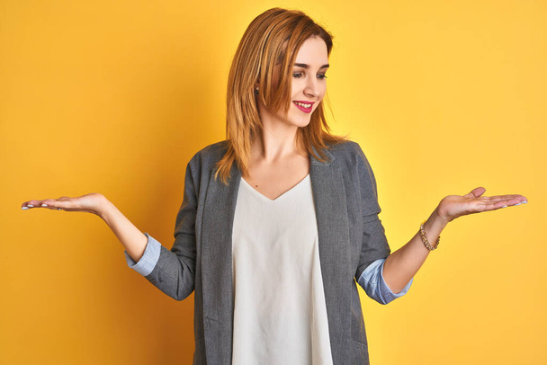 Rothaarige kaukasische Geschäftsfrau auf gelbem, isoliertem Hintergrund lächelnd mit beiden Händen geöffnete Handflächen, Präsentation und Werbung Vergleich und Ausgewogenheit - Foto, Bild