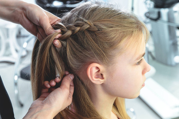Cabeleireiro tece uma trança para uma menina loira pré-adolescente em um salão de beleza e cabelo
 - Foto, Imagem