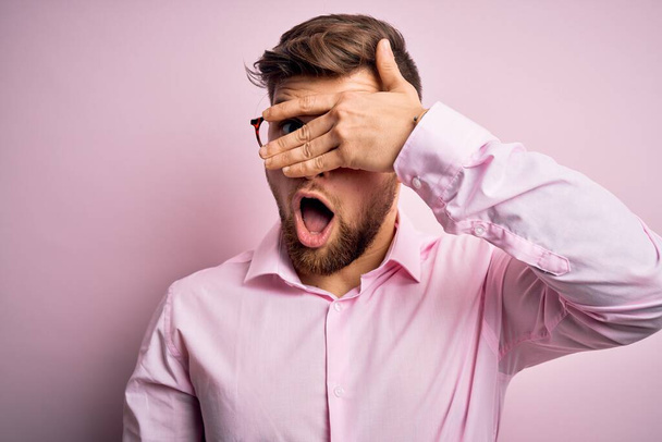 Young handsome blond man with beard and blue eyes wearing pink shirt and glasses peeking in shock covering face and eyes with hand, looking through fingers with embarrassed expression. - Photo, Image