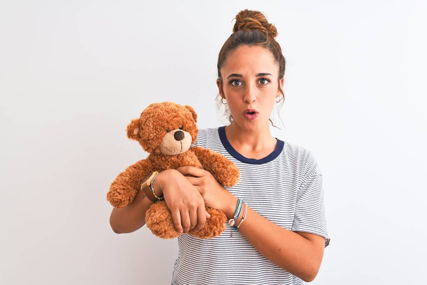 Young beautiful redhead woman holding stuffed teddy bear over isolated background scared in shock with a surprise face, afraid and excited with fear expression - Photo, image