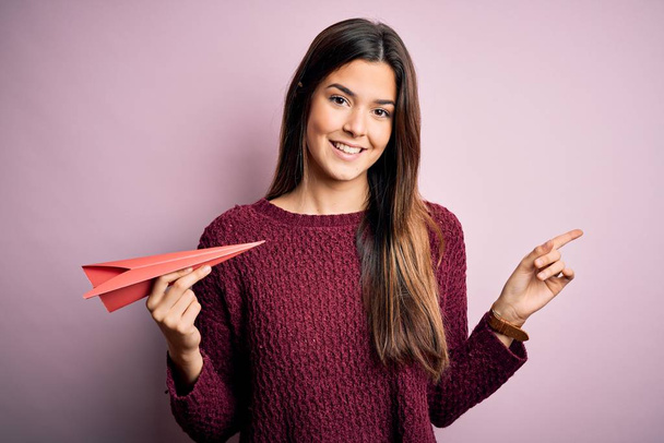 Young beautiful girl holding paper plane standing over isolated pink background very happy pointing with hand and finger to the side - Foto, Bild