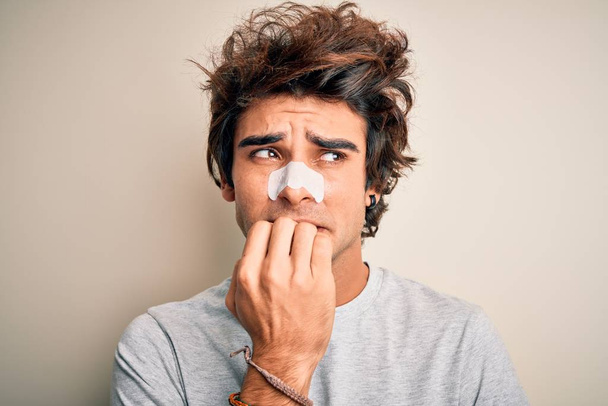 Νεαρός όμορφος άντρας που χρησιμοποιεί λουρίδα μύτης και στέκεται πάνω από απομονωμένο λευκό φόντο δείχνοντας αγχωμένος και νευρικός με τα χέρια στο στόμα να δαγκώνουν νύχια. Πρόβλημα άγχους. - Φωτογραφία, εικόνα