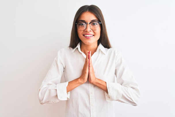 junge schöne Geschäftsfrau mit Brille steht vor isoliertem weißen Hintergrund und betet mit den Händen zusammen um Vergebung und lächelt selbstbewusst. - Foto, Bild