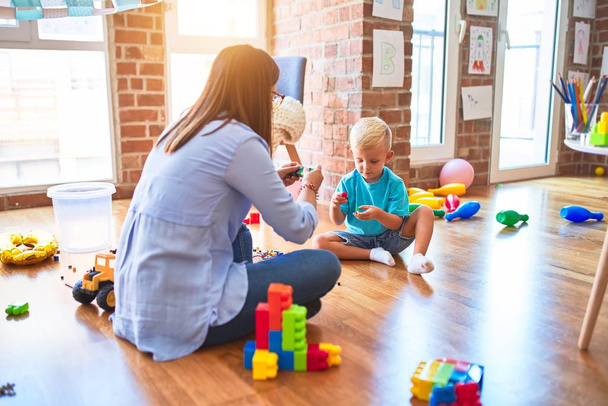 若い白人の子供は教師と一緒に学校で遊んでいる。母と息子-遊び場でおもちゃのブロックで塔を爆発 - 写真・画像